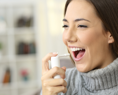 Happy woman wearing jersey using a analgesic spray to soften the throat sitting on a sofa in a house interior