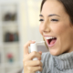 Happy woman wearing jersey using a analgesic spray to soften the throat sitting on a sofa in a house interior